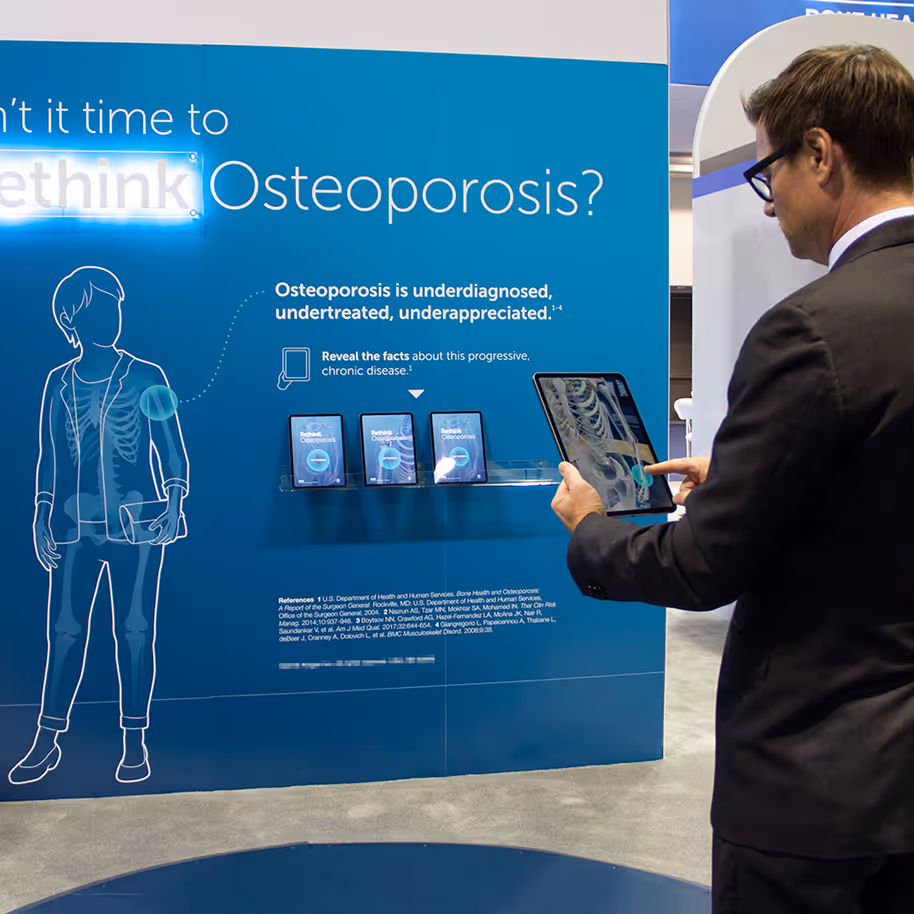 Man holding a tablet displaying a skeleton image, standing in front of an informational wall about osteoporosis underdiagnosis and treatment.