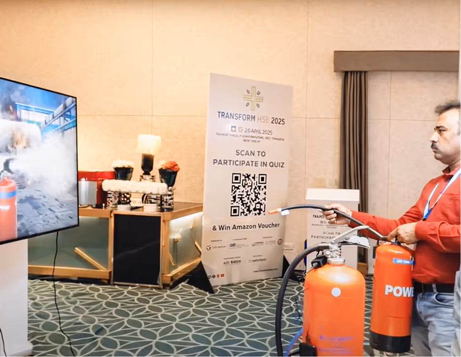 Man in red shirt demonstrating fire extinguisher usage in front of a screen showing fire safety visuals and a poster inviting participation in a safety quiz with QR code.