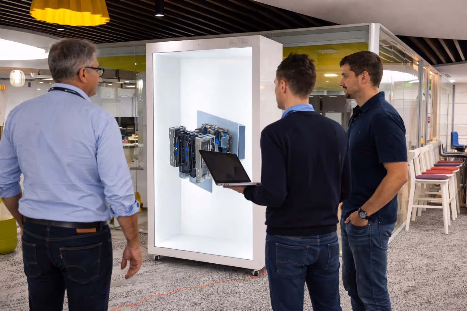 Three men standing in an office space observing a mounted display of computer hardware with one man holding a laptop.