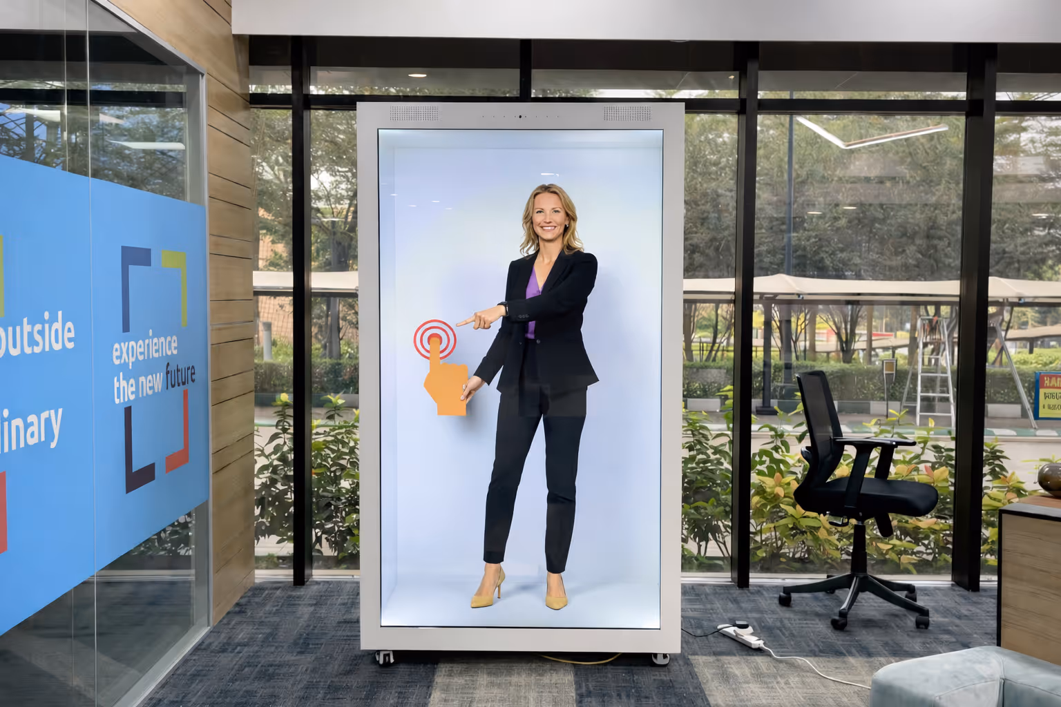 A woman in business attire smiling and pointing at a large interactive digital touchscreen inside a modern office.
