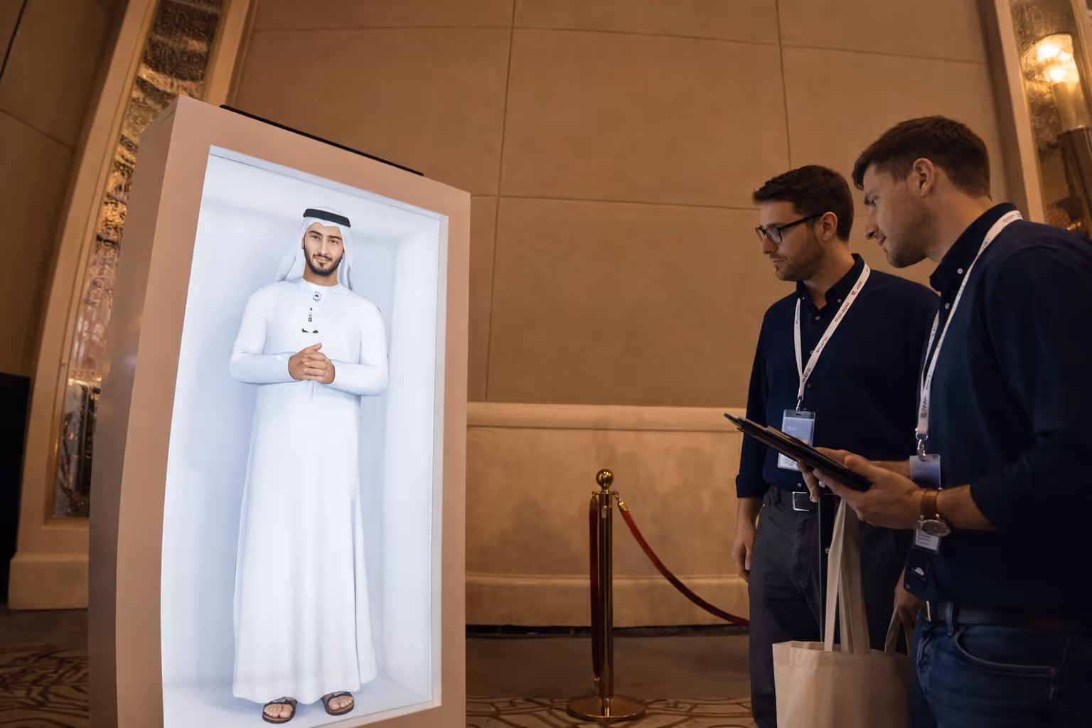 Two men with conference badges observing a digital display of a man in traditional Middle Eastern attire.