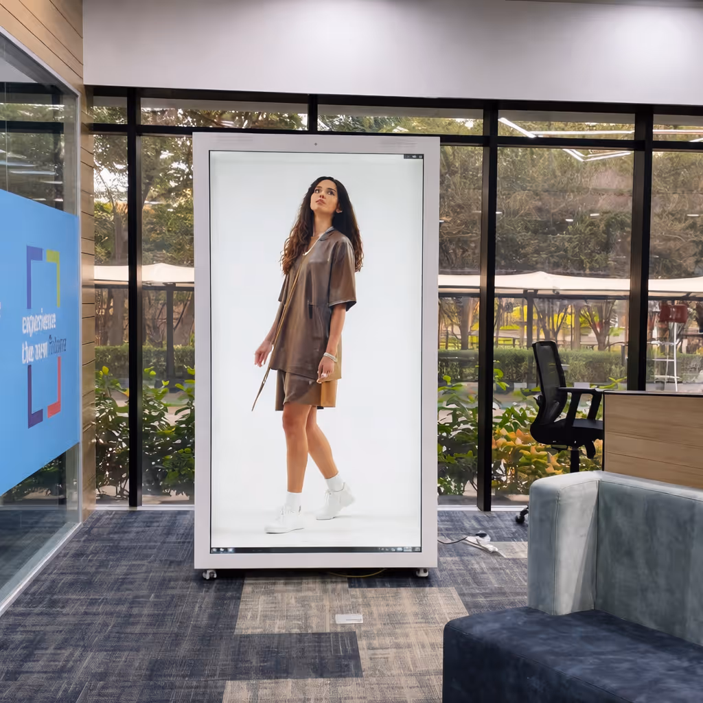 Indoor office space with carpeted floor and glass windows showing greenery outside, featuring a large vertical digital display of a woman wearing a brown outfit and white sneakers.