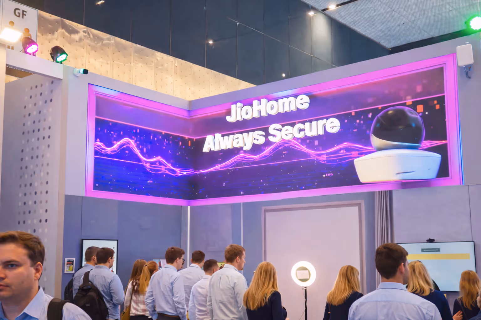 Group of people standing and facing a large screen displaying the text 'JioHome Always Secure' with a security camera image.
