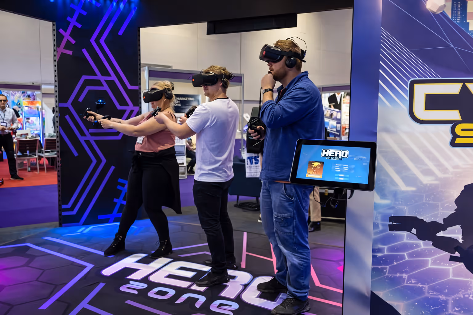 Three people standing in a VR gaming booth wearing headsets and holding controllers, playing a game called Hero Zone.