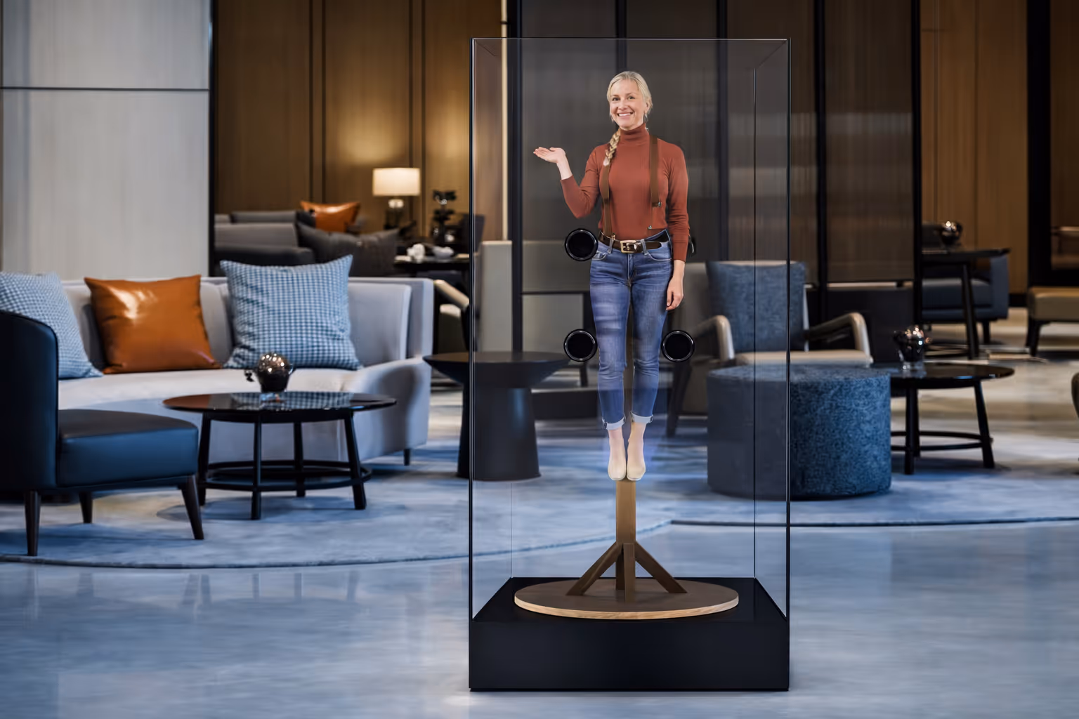 A woman suspended inside a glass display case in a modern lounge area with sofas and tables.