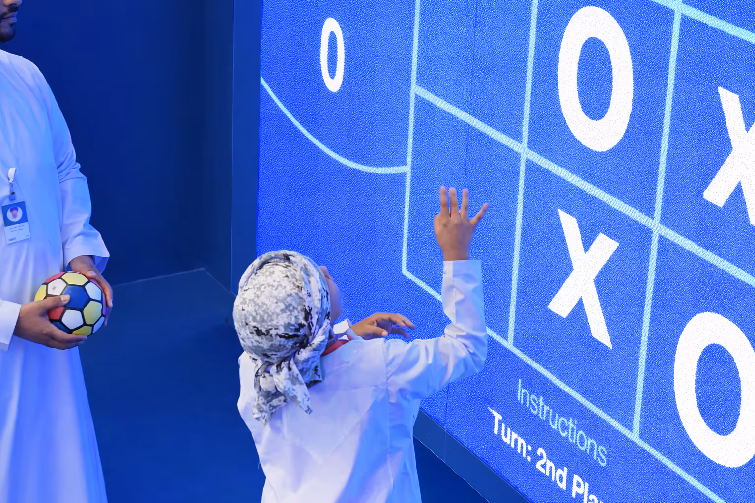 Child wearing traditional clothing playing a large digital tic-tac-toe game on a screen, with an adult holding a colourful soccer ball watching nearby.