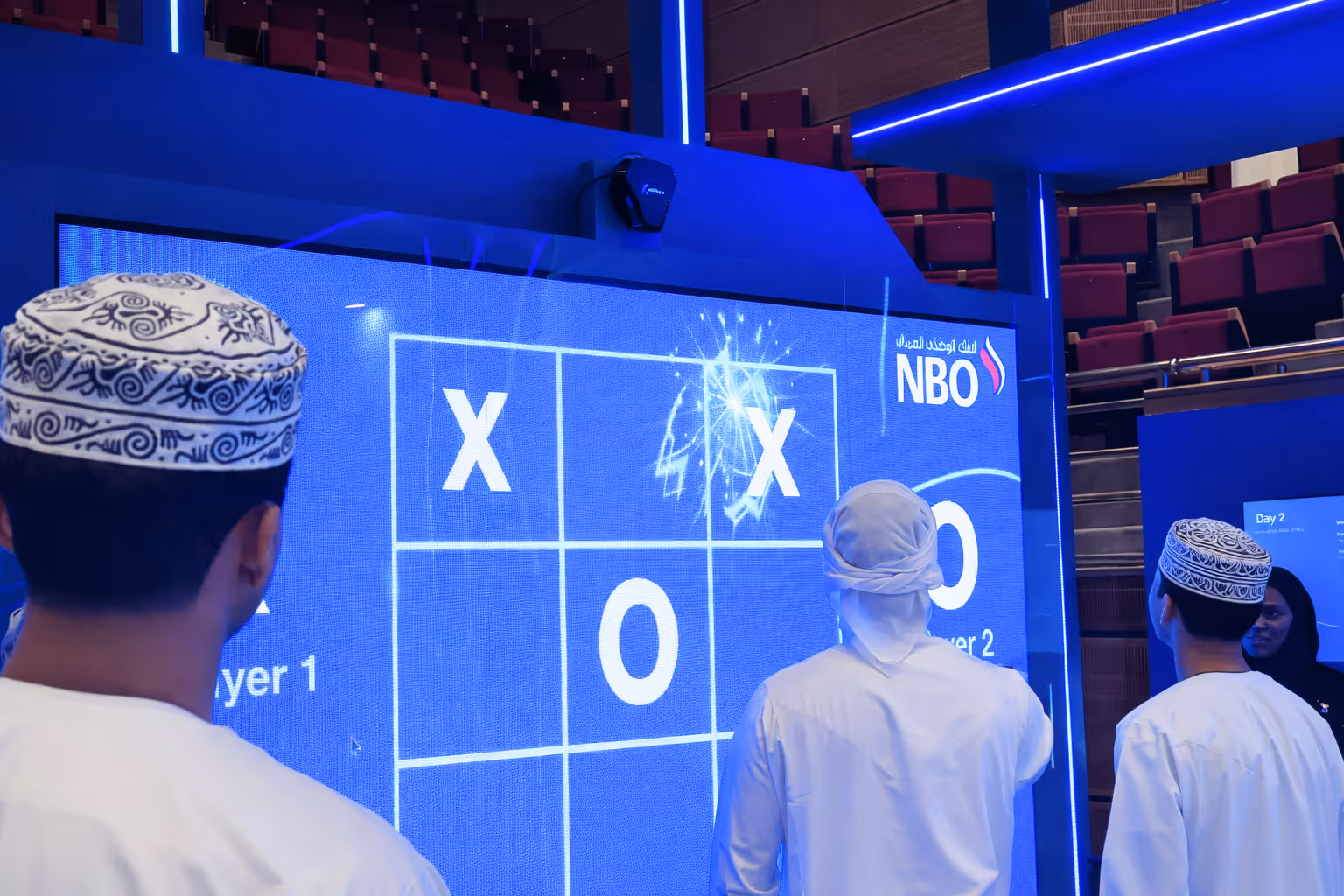 Three men in traditional Omani attire playing tic-tac-toe on a large digital screen showing the game in progress with X and O symbols.