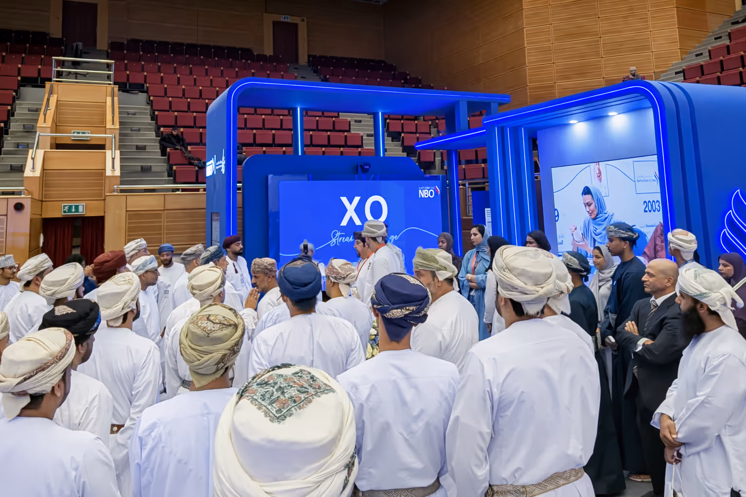 Group of people in traditional Middle Eastern attire gathered around a blue exhibition booth with 'XO' and NBO branding in an indoor auditorium.