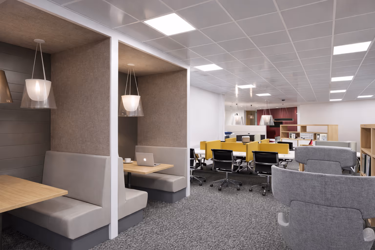 Modern office space with grey booth seating on the left, yellow divider panels with black office chairs in the centre, and grey cushioned chairs on the right.
