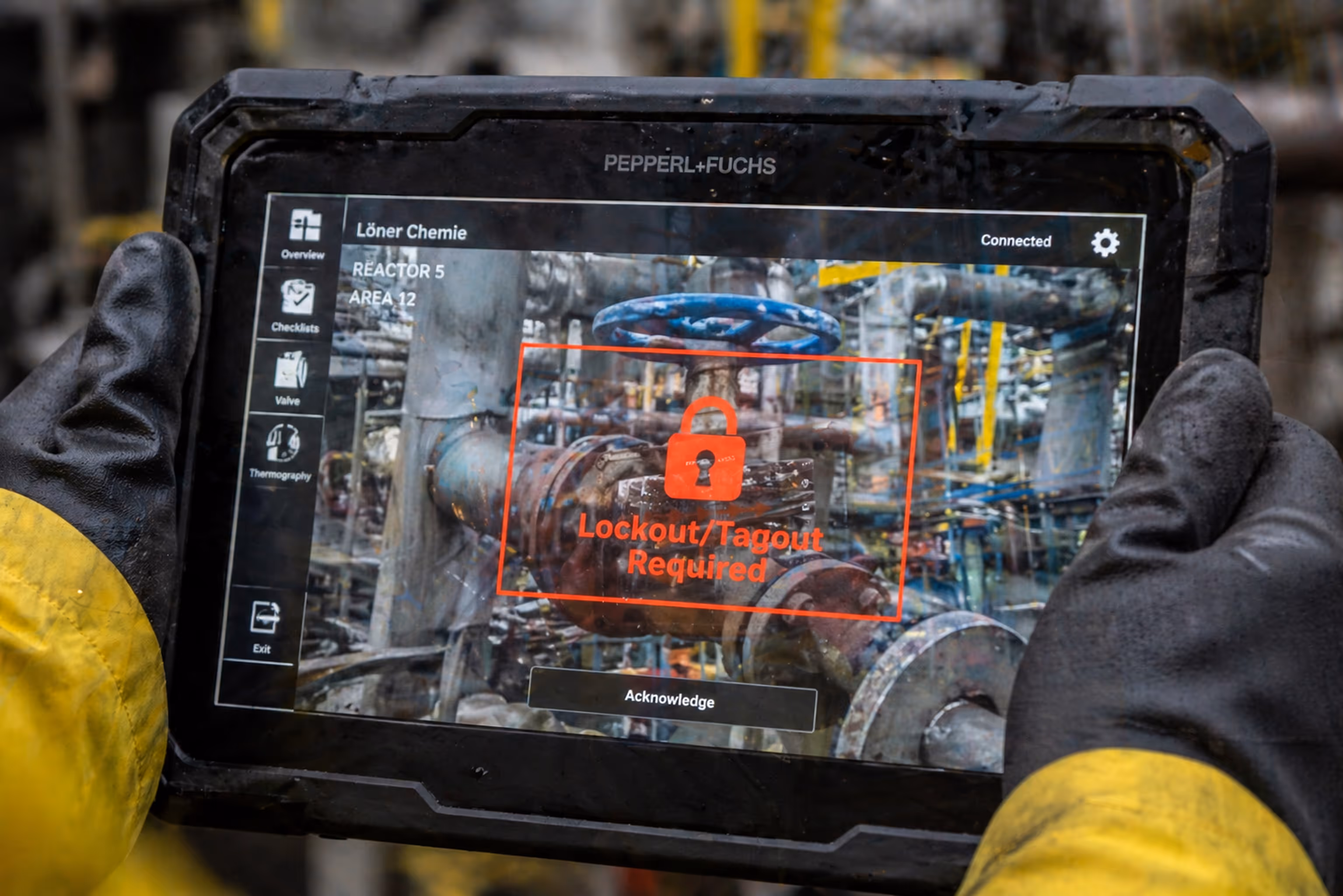 Industrial worker wearing gloves holding a rugged tablet displaying a red lock warning with 'Lockout/Tagout Required' over a valve in a chemical plant.