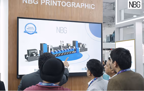 Group of people observing and discussing a large screen displaying a digital graphic of industrial printing machinery at NBG Printographic booth.