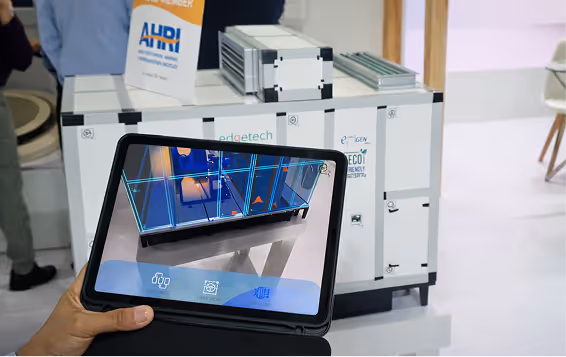 Hand holding a tablet displaying a 3D digital model of an industrial machine in front of the actual machine.