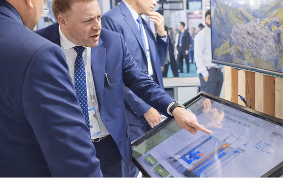 Business professionals in suits interacting with a large touchscreen display showing a technical schematic at a trade show or conference.