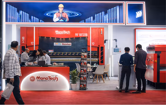 Trade show booth for MonoTech Engineers Pvt. Ltd. featuring a large industrial machine, a reception desk, brochure stand, and attendees interacting.