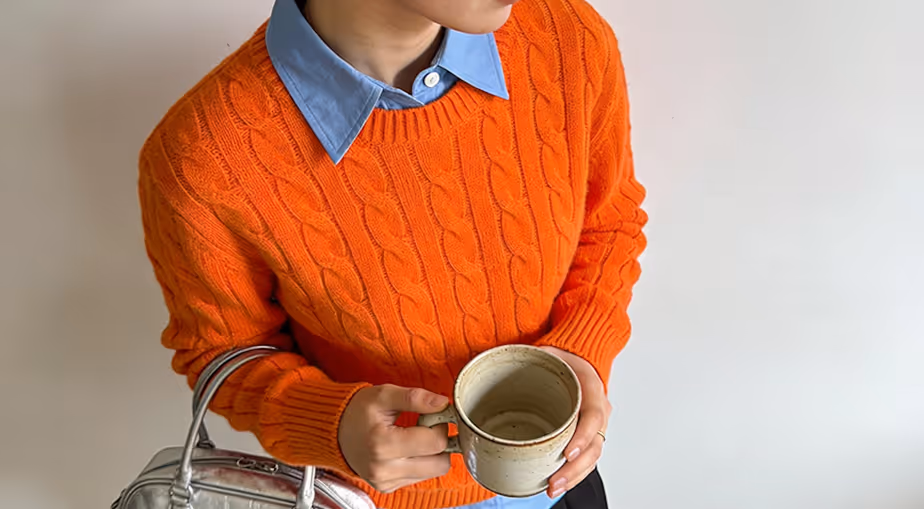 Person wearing an orange cable-knit sweater over a blue collared shirt holding a ceramic mug and a silver handbag.