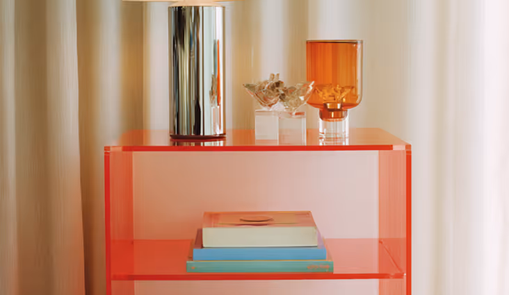 Red translucent side table with stacked books on the lower shelf and decorative glass pieces and a metal vase on top, set against light curtains.