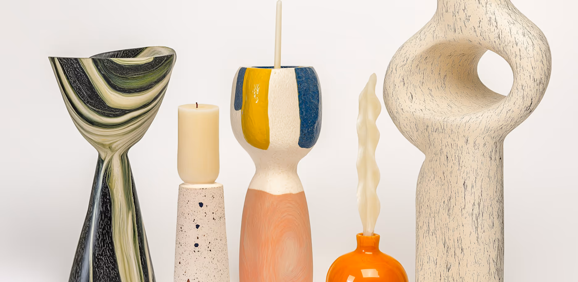 Five decorative candles in uniquely shaped holders including marbled black and green, speckled beige, multicolor, orange, and textured cream designs.