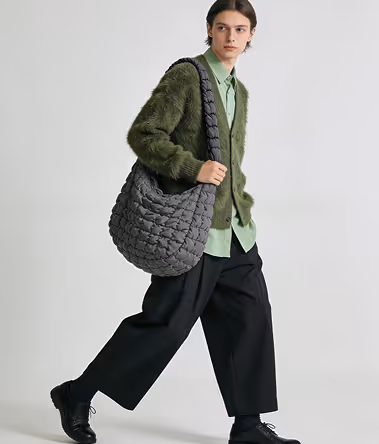 Young person wearing a green fuzzy cardigan over a light green shirt, black wide-leg pants, black shoes, and carrying a large gray quilted shoulder bag.