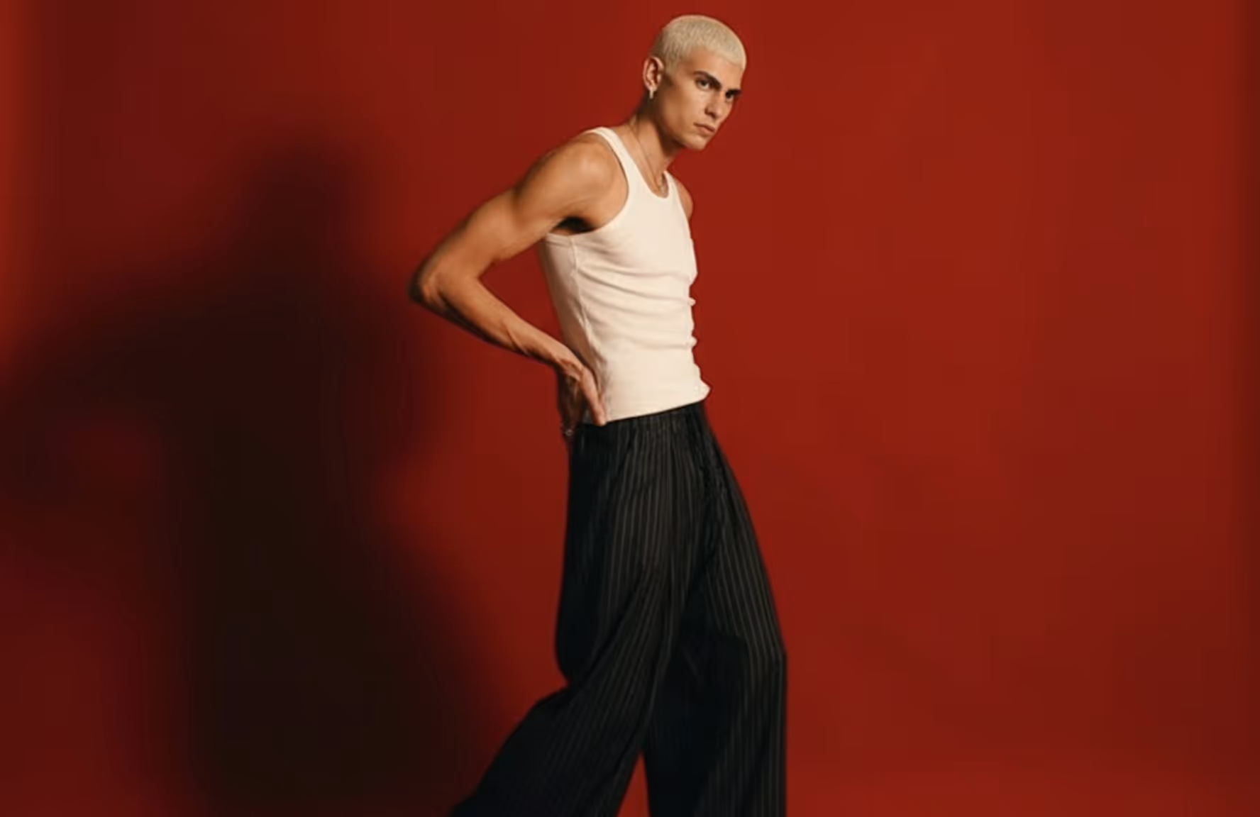 Young man with platinum blonde hair wearing a white tank top and black striped wide-leg pants against a red background.