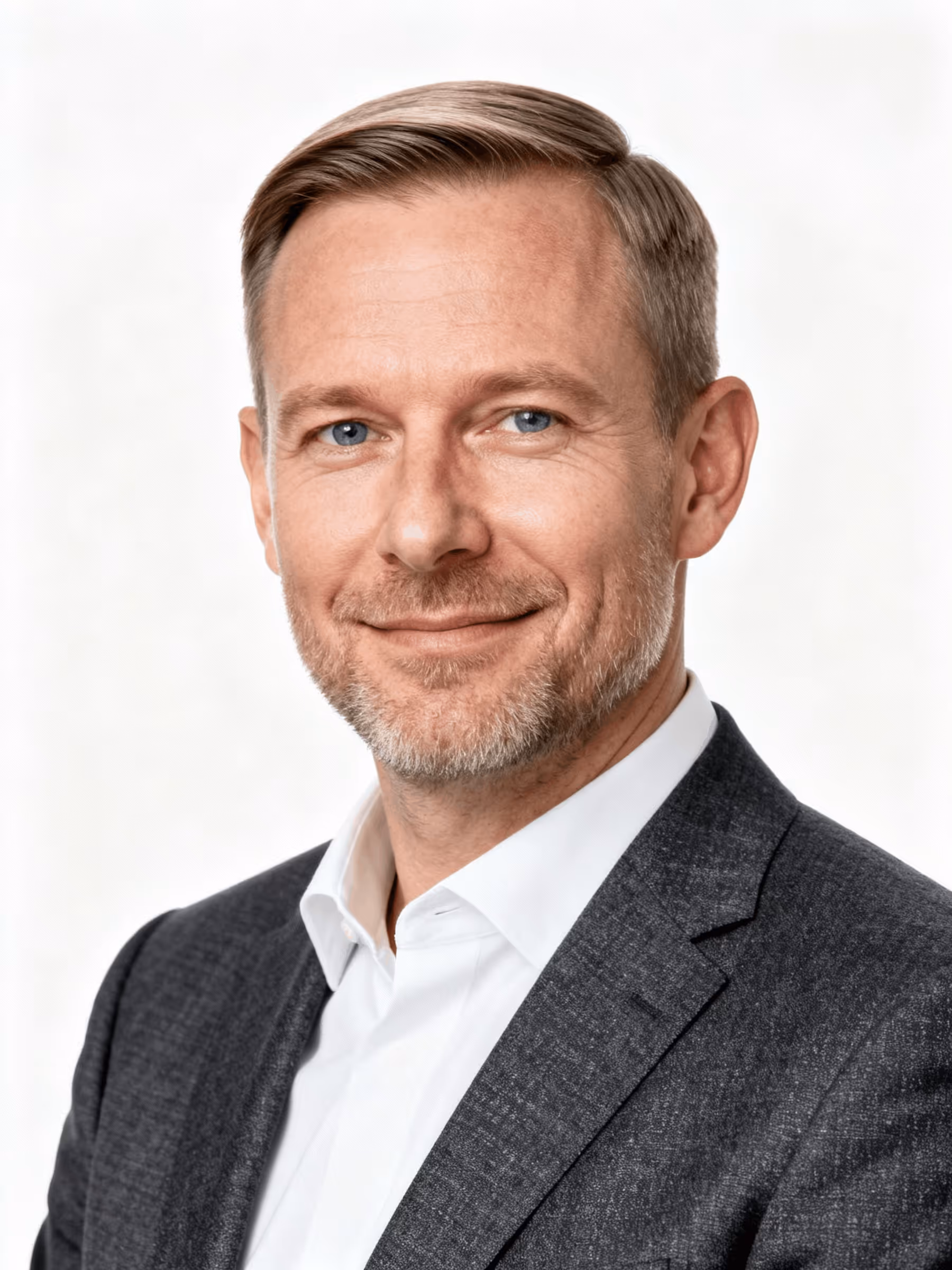 Middle-aged man with short light brown hair and blue eyes wearing a dark blazer and white shirt smiling gently against white background.