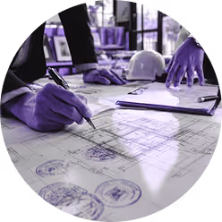 Person wearing gloves drawing on architectural blueprints with a pen, with a clipboard and white hard hat nearby.