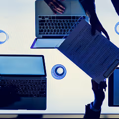 Overhead view of a desk with two laptops, a tablet, and hands holding a clipboard with a document.