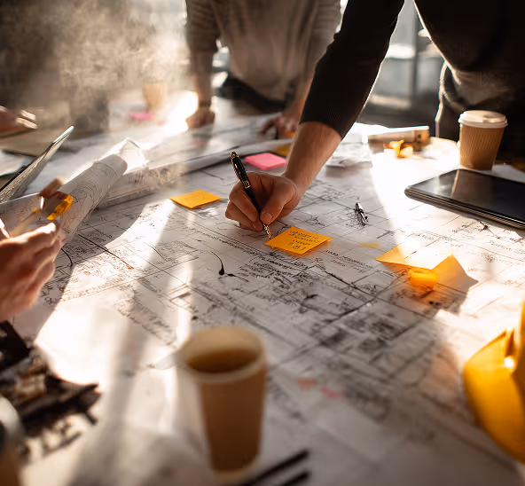 People reviewing architectural blueprints on a table with sticky notes, a tablet, and coffee cups in a bright workspace.