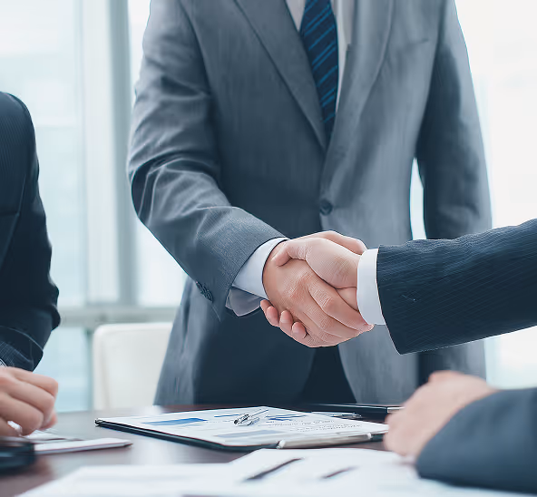 Two business professionals shaking hands over a desk with documents and a pen, signifying agreement or partnership.