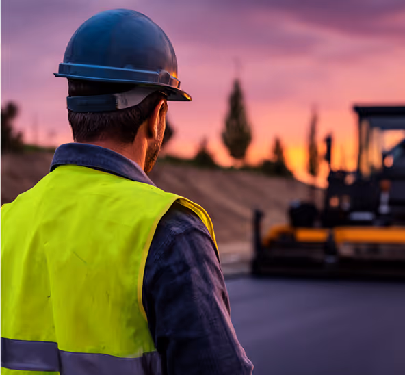 Construction worker in safety helmet and yellow vest overseeing asphalt paving at sunset.
