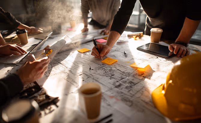 People reviewing architectural blueprints on a table with sticky notes, a tablet, and coffee cups in a bright workspace.