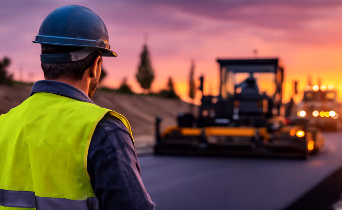 Construction worker in safety helmet and yellow vest overseeing asphalt paving at sunset.