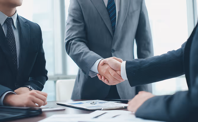 Two business professionals shaking hands over a desk with documents and a pen, signifying agreement or partnership.