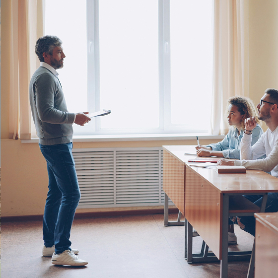Man standing and speaking to two students sitting at desks in a classroom with a large window.