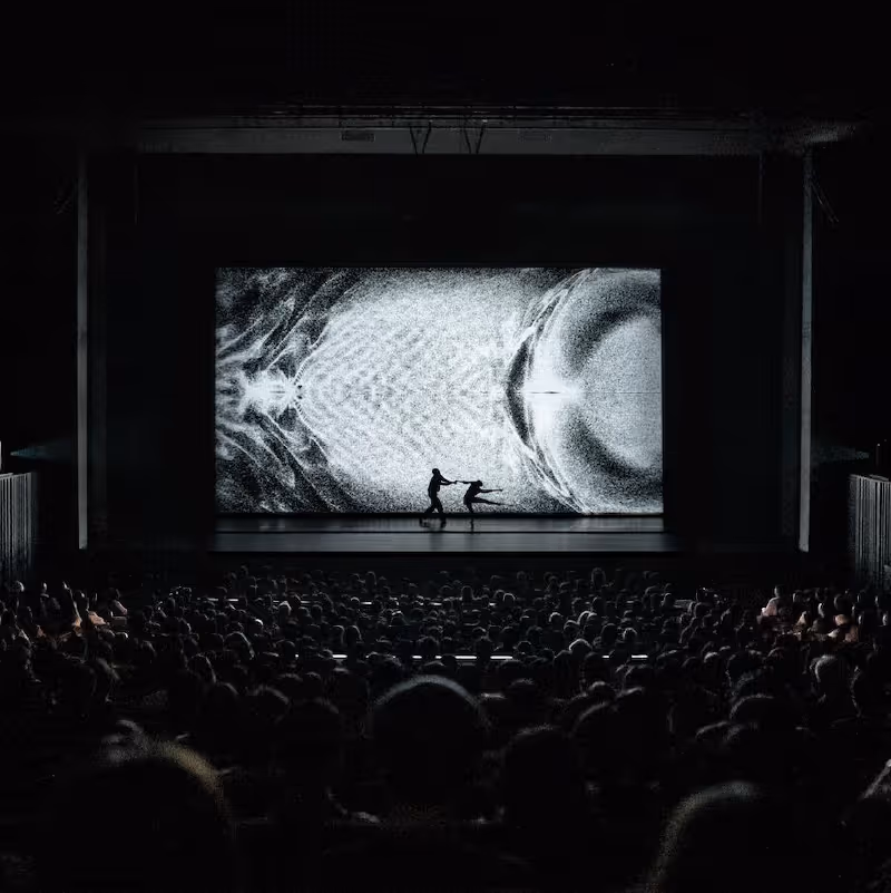 Deux danseurs sur scène avec un grand écran abstrait noir et blanc derrière, public en avant.
