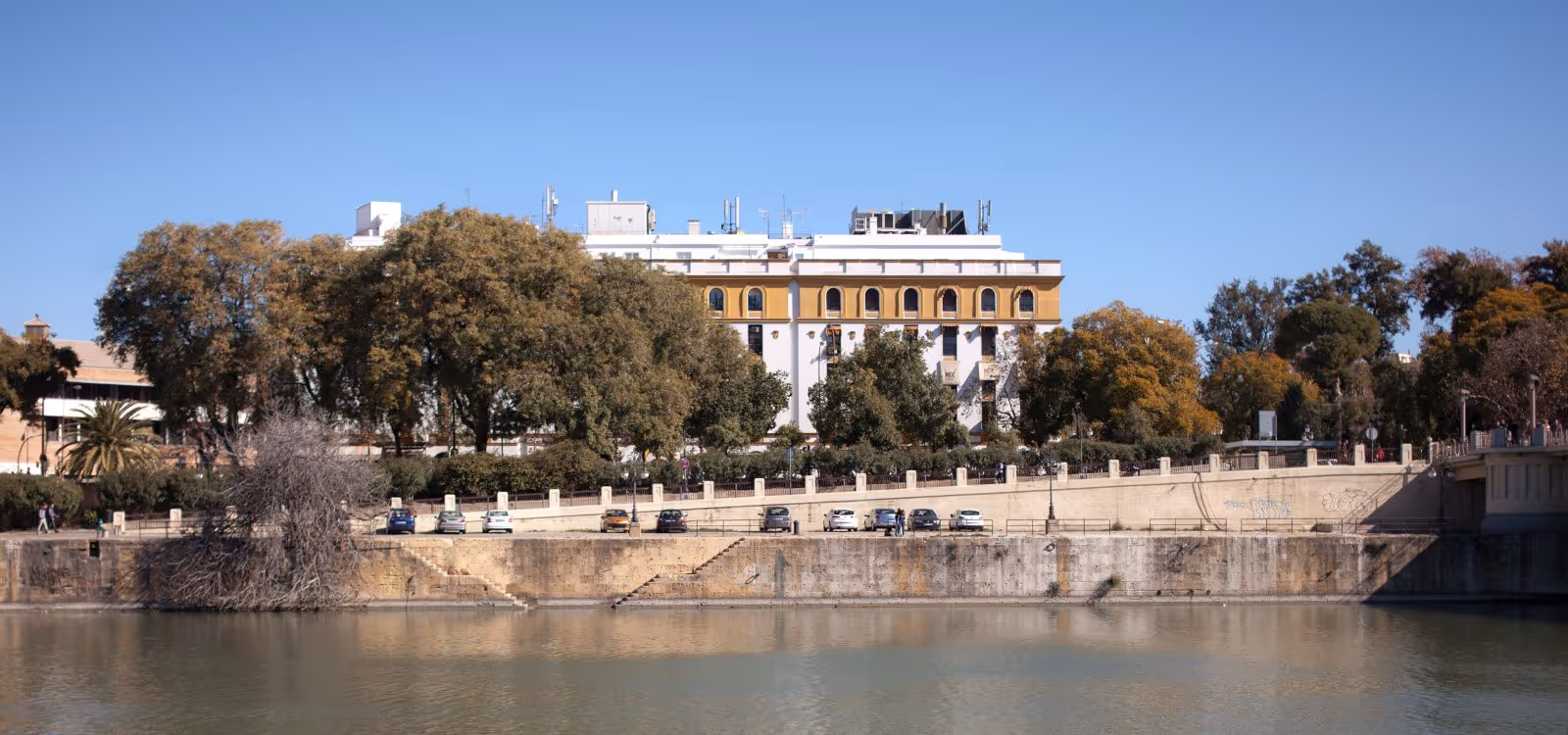 Riverside view of a classical ochre and white building lined with trees, seen across calm water from an embankment with parked cars