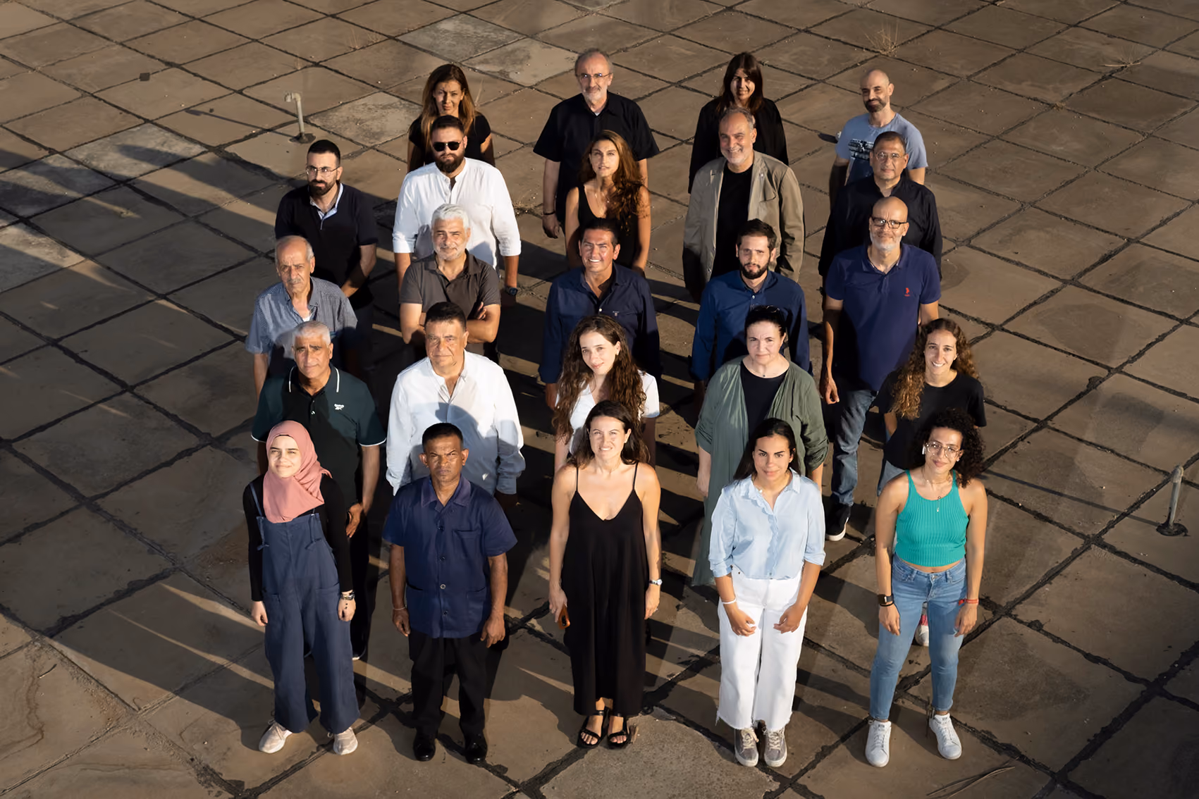 Overhead view of a large team gathered in rows on a paved outdoor terrace, casting long shadows in warm afternoon light