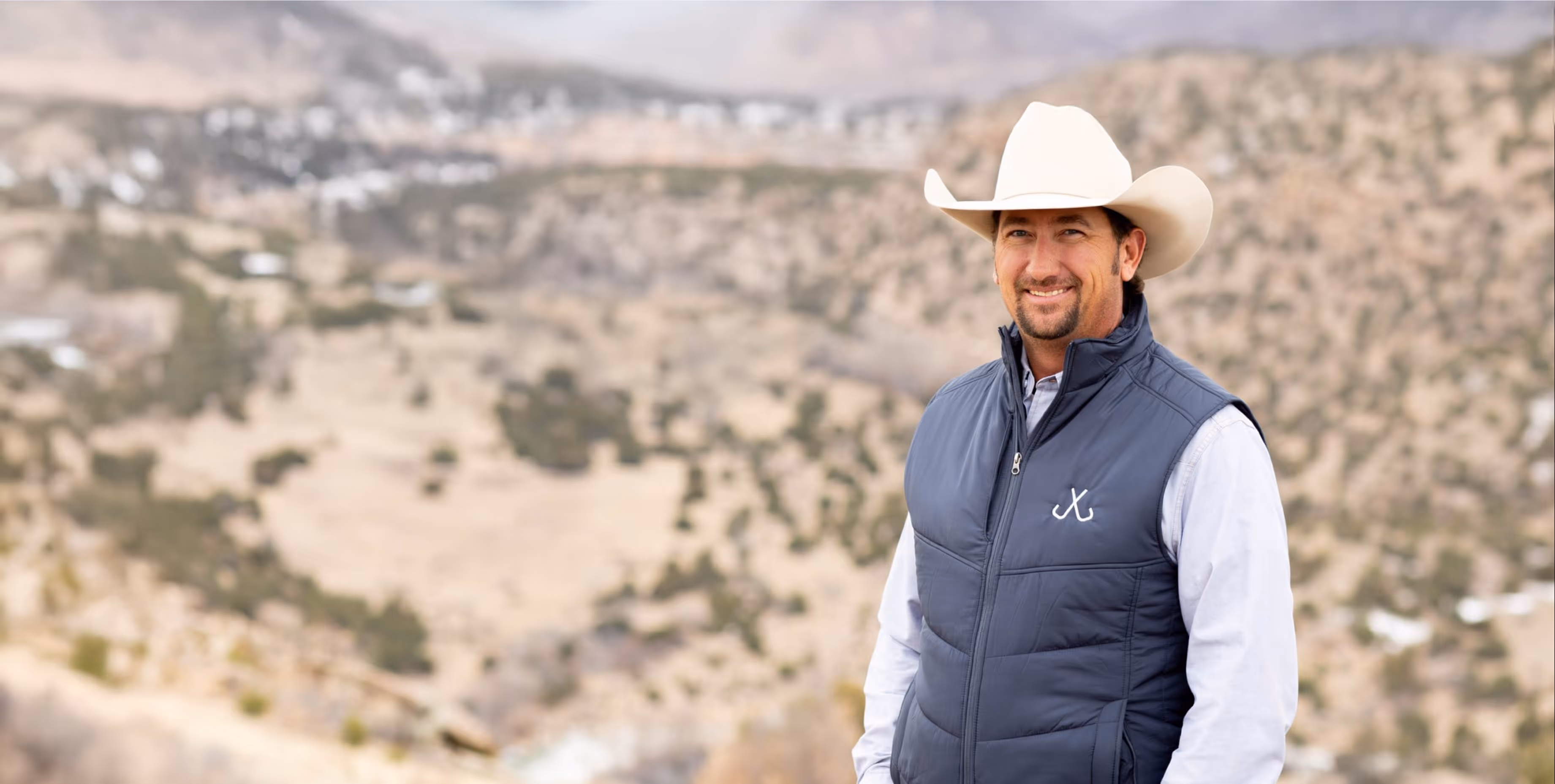 Man wearing a white cowboy hat and navy vest standing outdoors with a blurred mountainous landscape in the background.