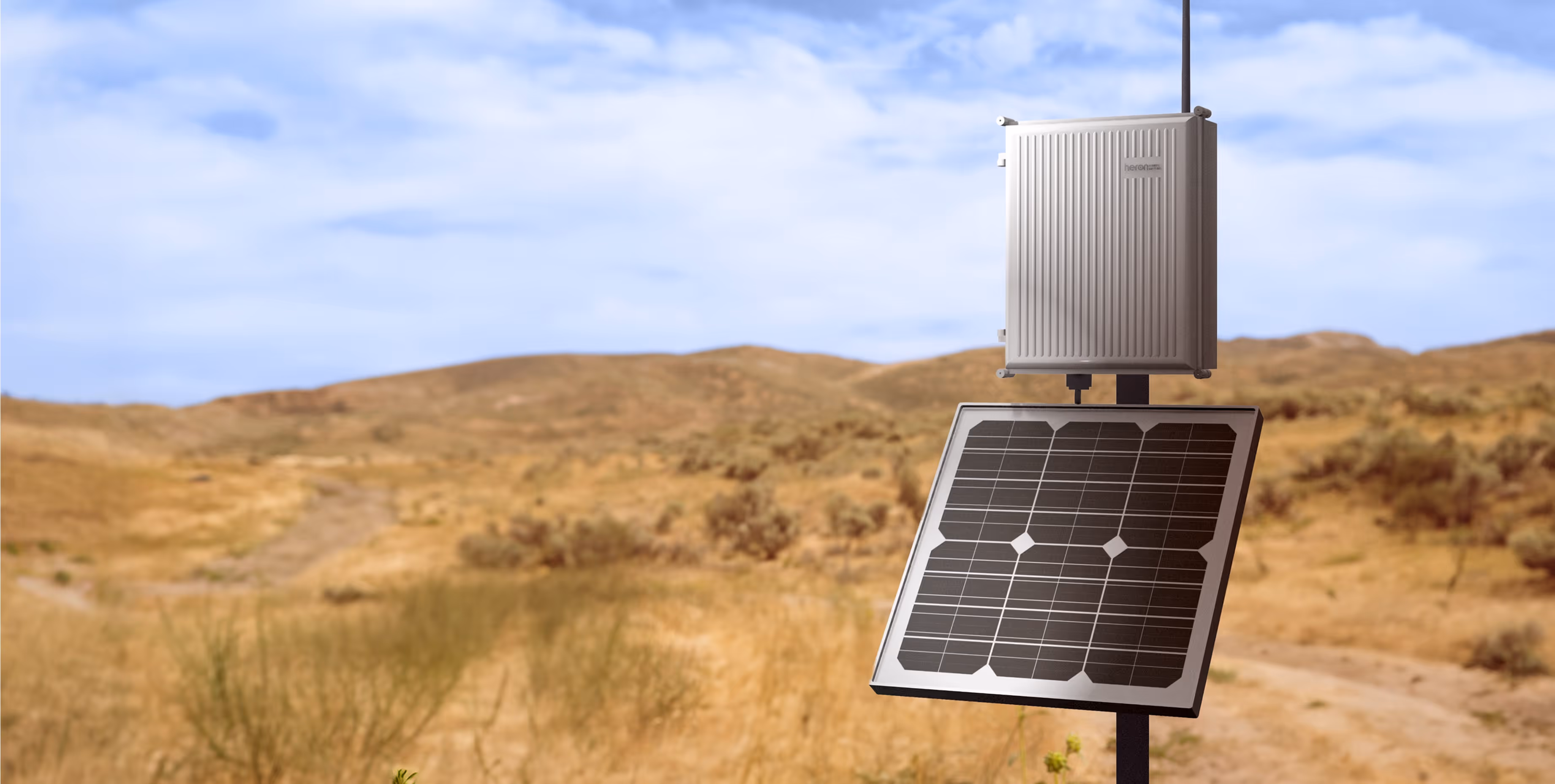 Small solar panel mounted on a pole with a weatherproof electronic box attached above it in a dry, hilly landscape under a blue sky.