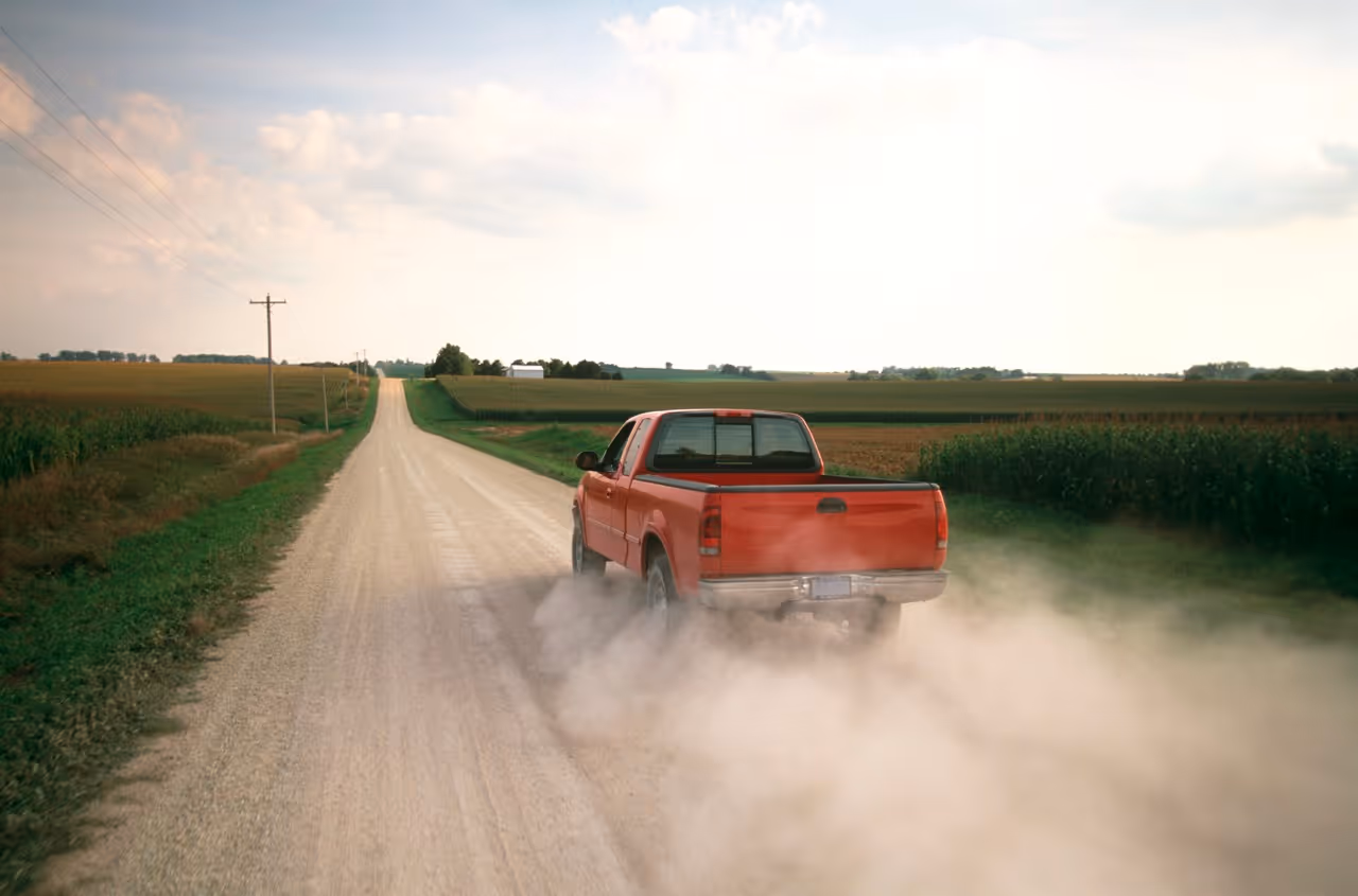 Red pickup truck driving away on a rural dirt road, kicking up dust with fields and power lines on either side.