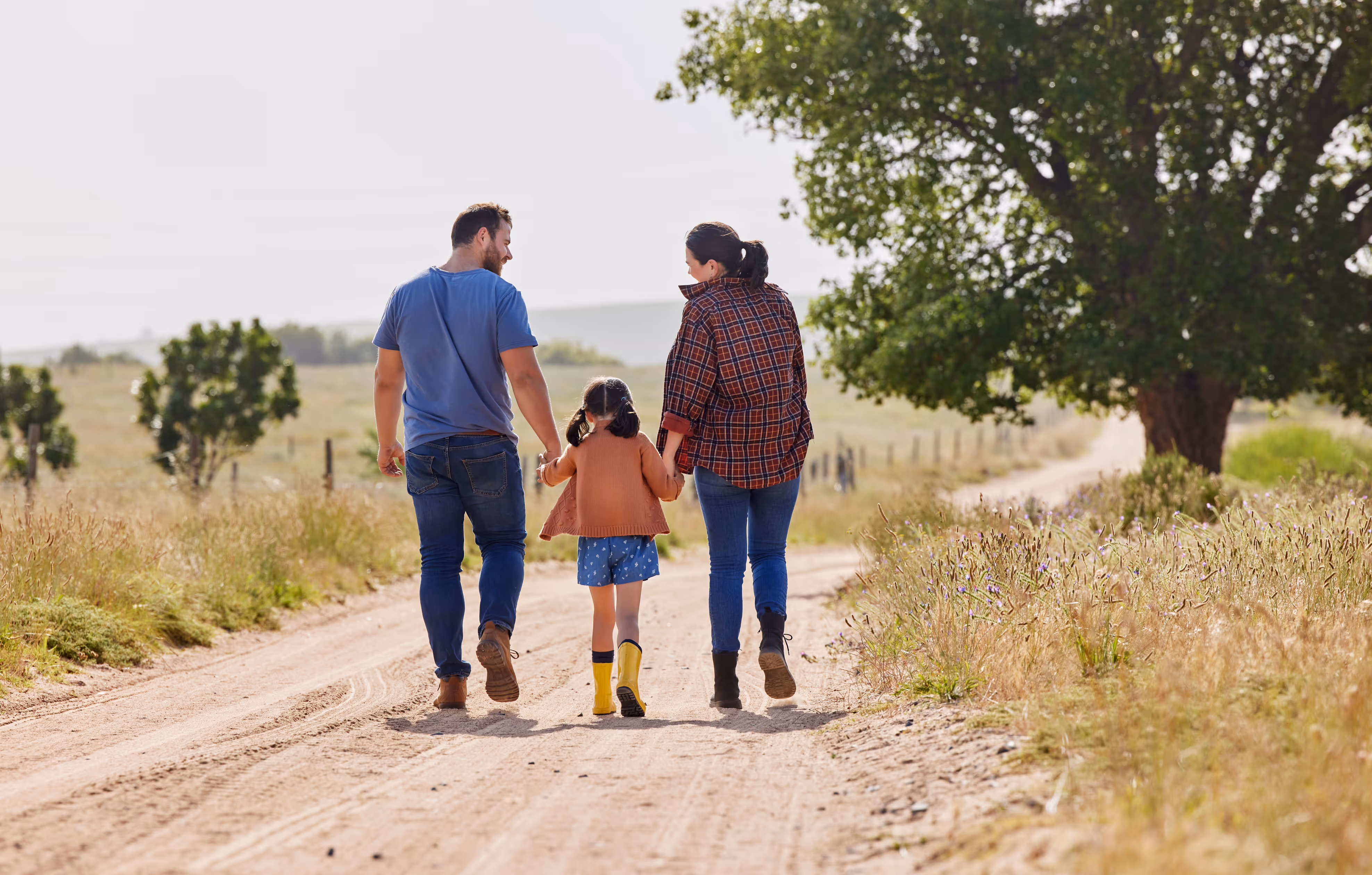 A man, woman, and child holding hands walking down a dirt country road surrounded by grass and trees.