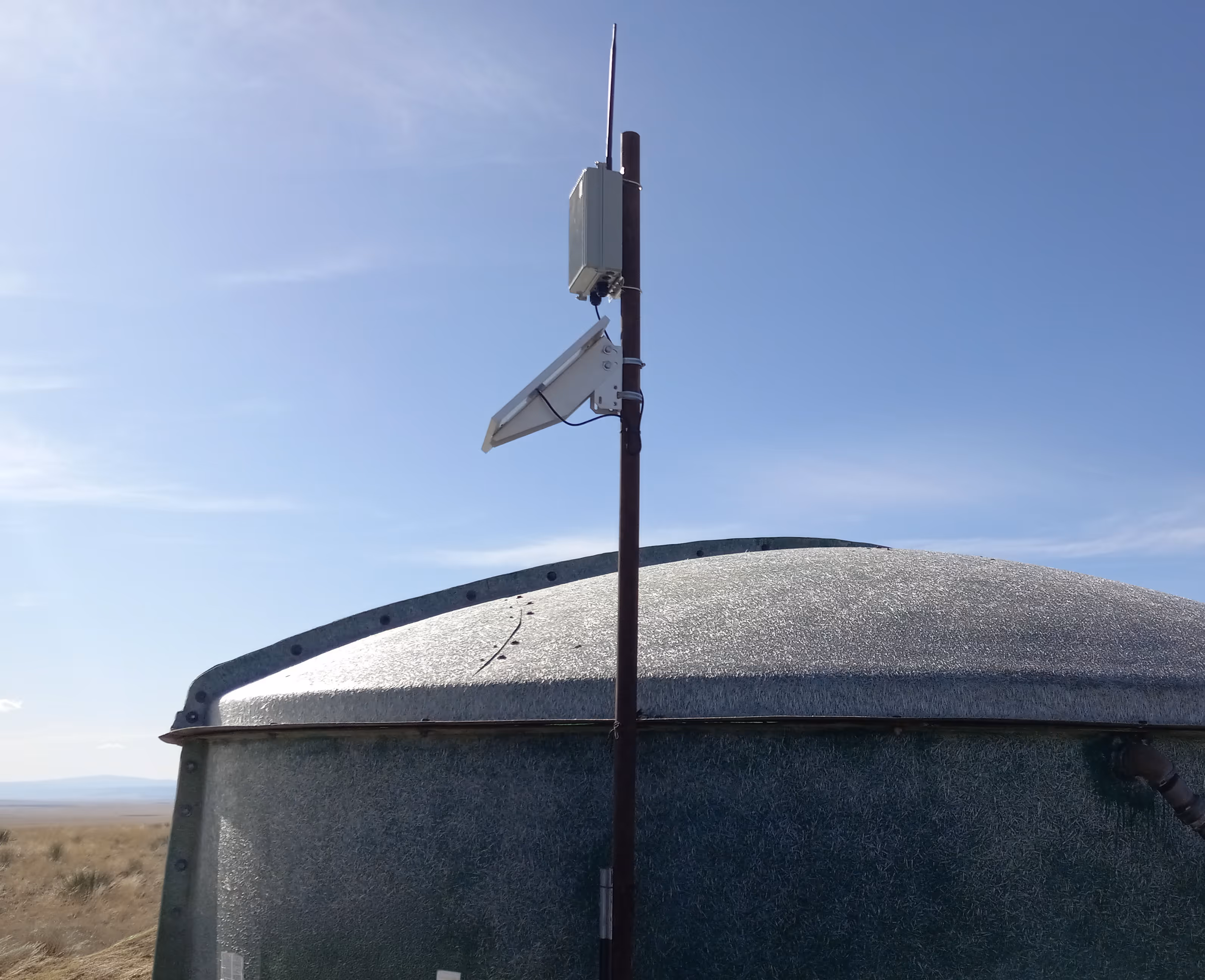 Metal storage tank with a mounted pole supporting a small solar panel and a white box under a clear blue sky.