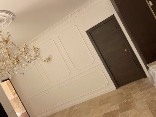 Interior view of a room with beige tiled floor, cream-colored wall panels, a dark wooden door, and an ornate crystal chandelier.