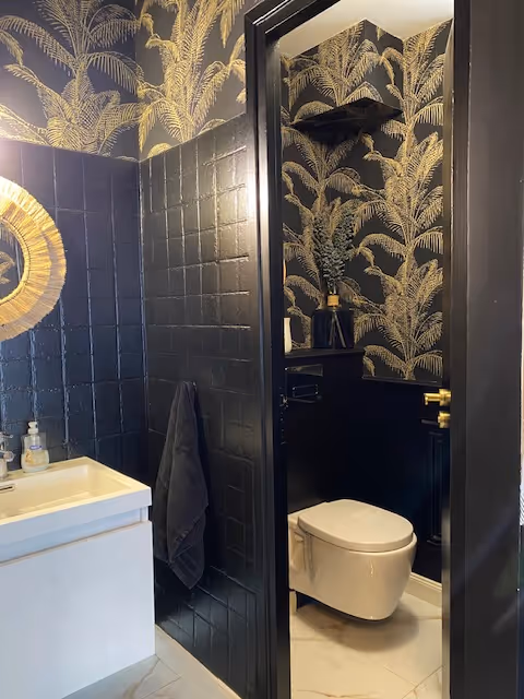 Modern bathroom with black tiled walls, gold palm leaf wallpaper, white floating toilet, white sink, and a round mirror with a golden frame.