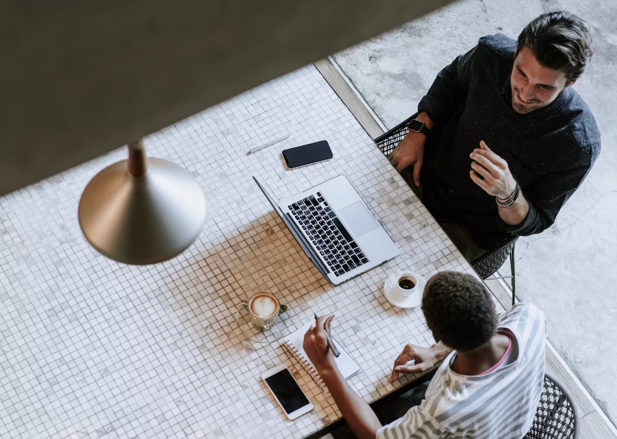 Two people at a tiled table with a laptop, smartphones, coffee, and a notebook in a casual meeting.