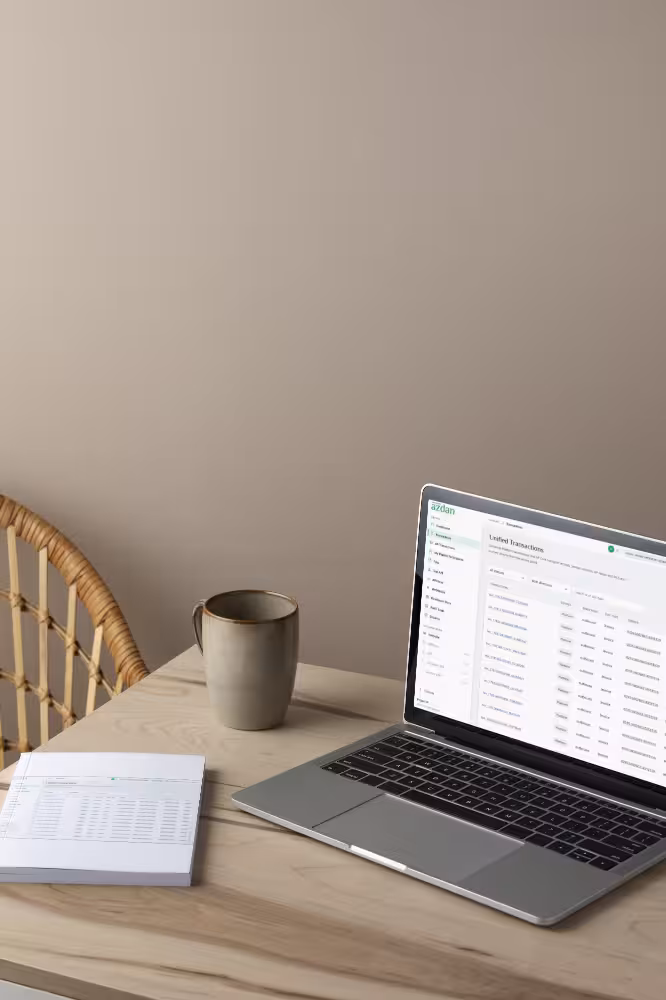 Laptop showing transactions, mug, and closed notebook on a light wooden table with a rattan chair.