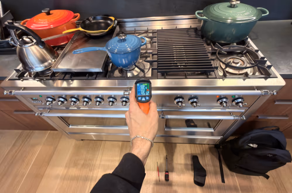 Person holding an infrared thermometer aimed at a stainless steel stove with various pots and pans on the burners.