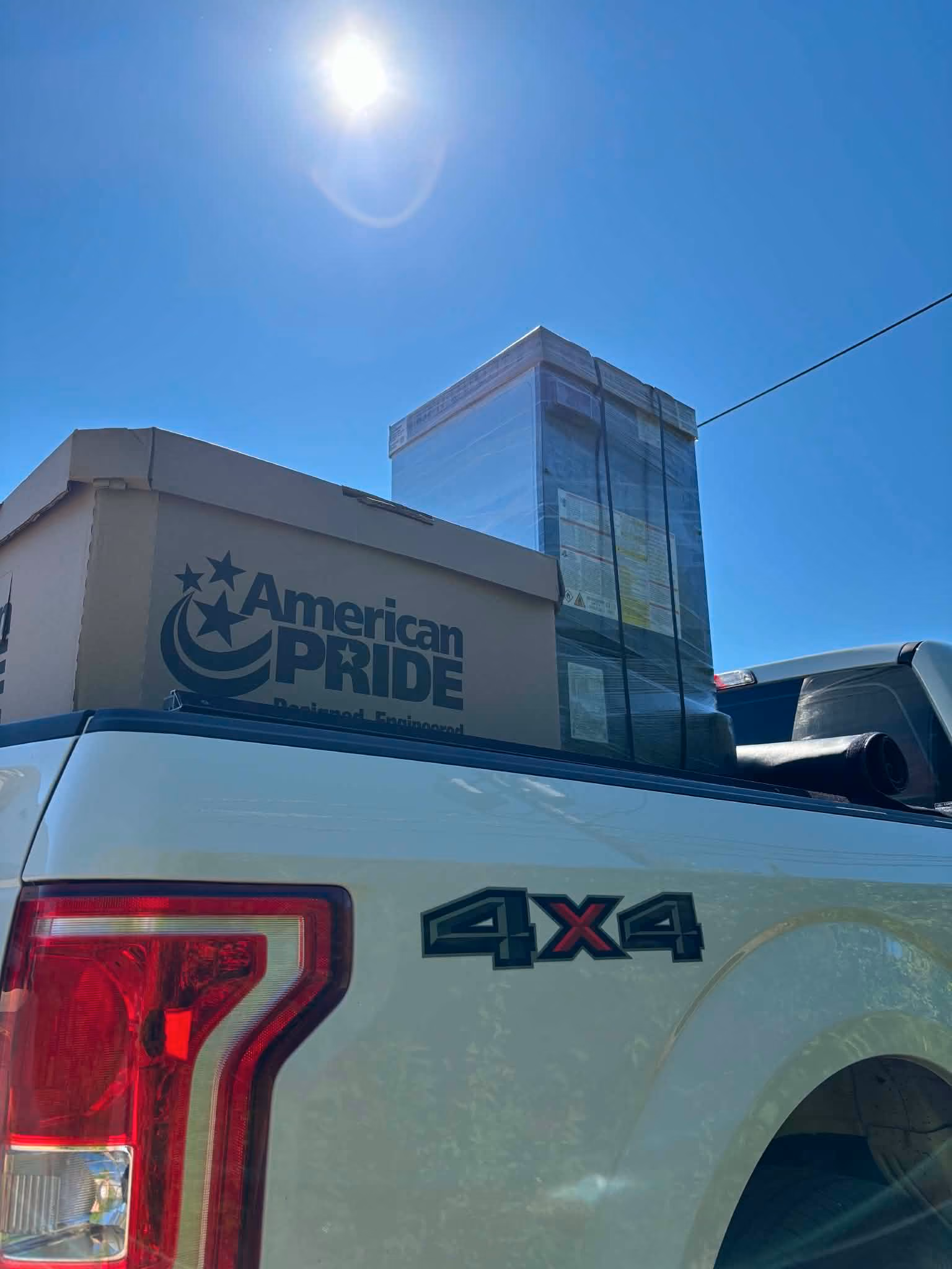 White 4x4 truck with American Pride HVAC equipment boxes in the bed under bright sun.
