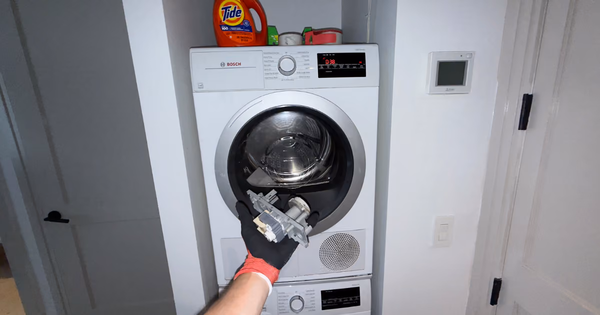 Gloved hand holding a removed component in front of a stacked dryer and washer.