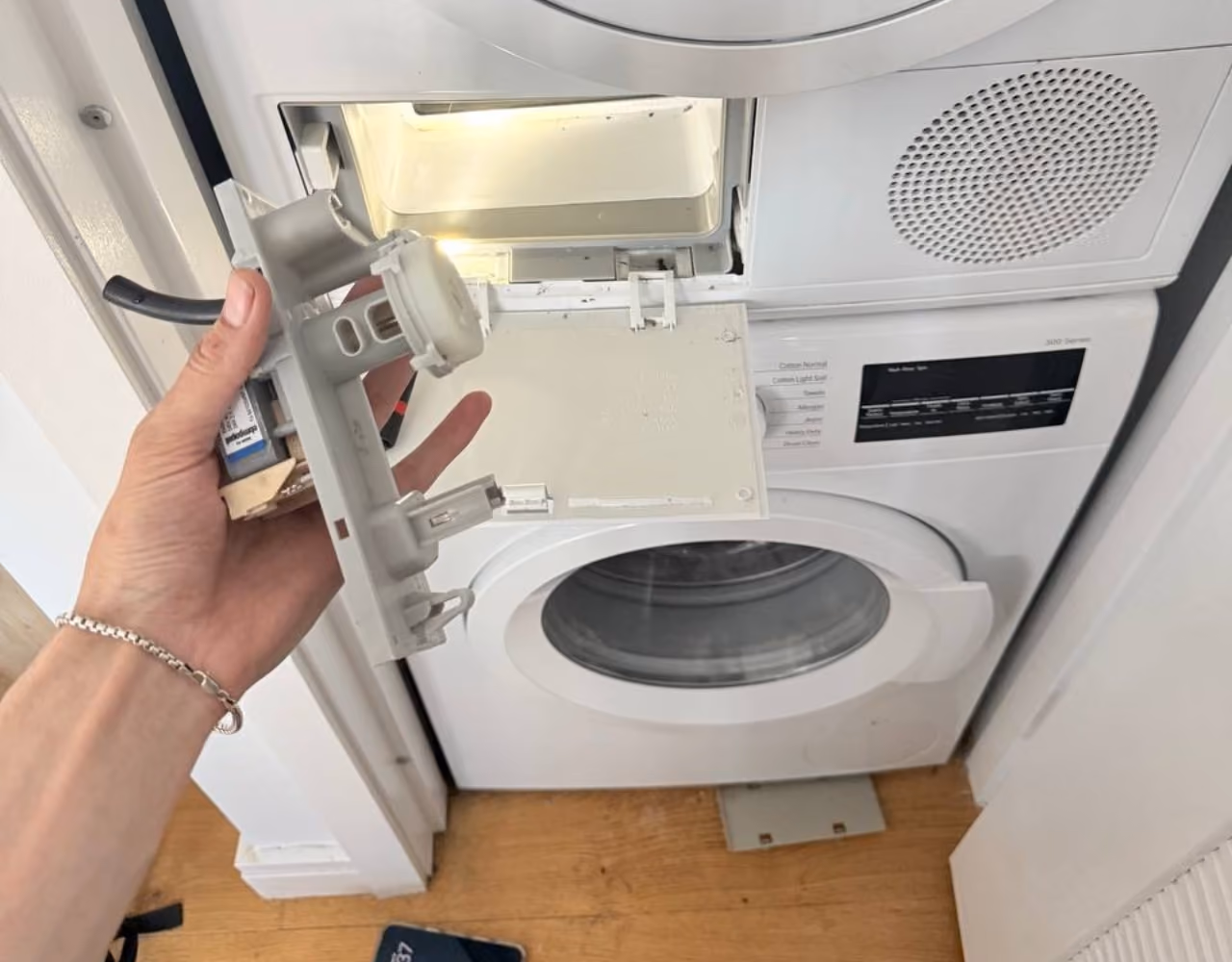 Hand holding a removed pump assembly beside an open dryer service compartment.