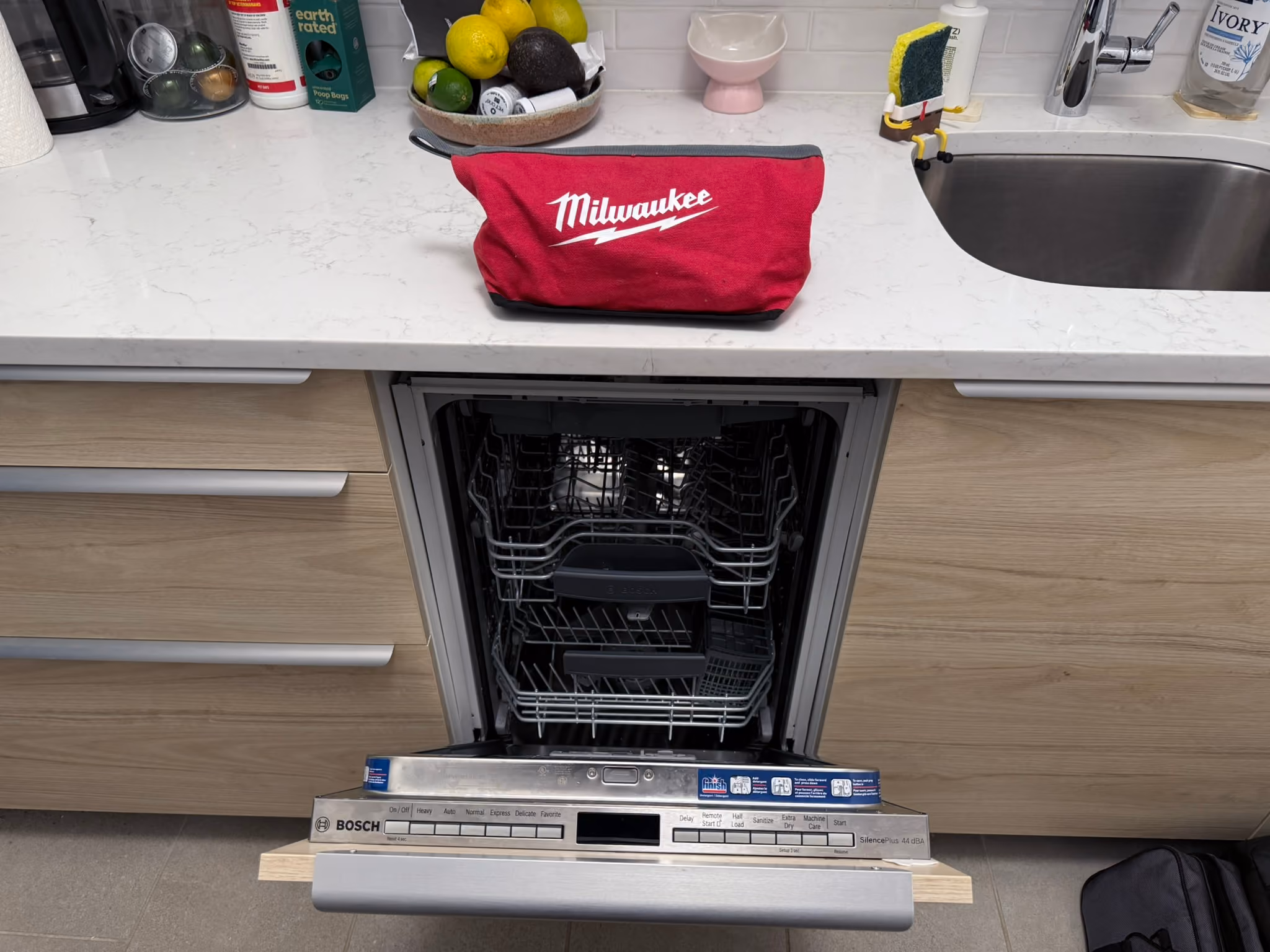 Open dishwasher beneath a kitchen counter with a red tool bag above it.